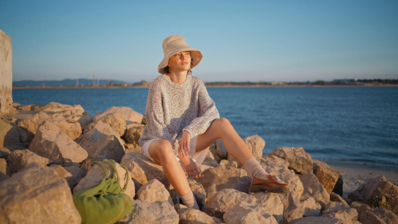 Žena sedi na stenama pored mora u ležernom outfitu sa šeširom, pletenim džemperom i smeđim brodaricama.