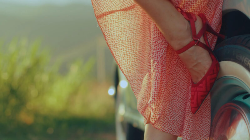 Ženska noga u crvenim cipelama i prolećnoj haljini, detalj u retro stilu pored automobila, na suncem okupanom putu.