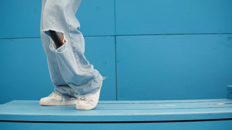 Bele retro patike i široke, pocepane farmerke u svetlom denimu na plavoj drvenoj klupi, street style estetika.