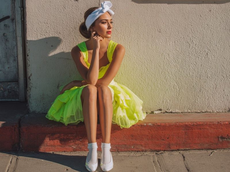  Devojka u jarko žutoj fluorescentnoj haljini, sa belim baletankama i belom trakom za glavu, sedi na trotoaru pored bele zida i crvenog ivičnjaka.