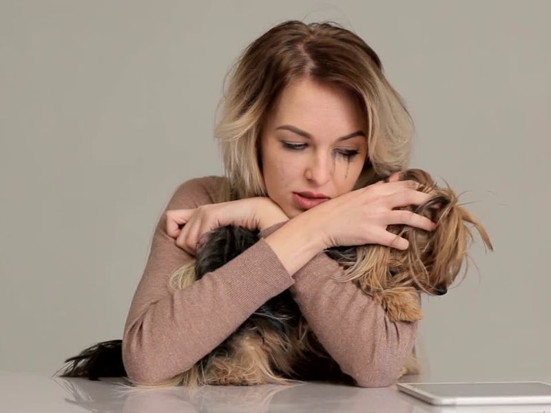  Tužna žena sa razmazanom maskarom grli malog psa dok sedi za stolom, izražavajući emotivnu povezanost i tugu.